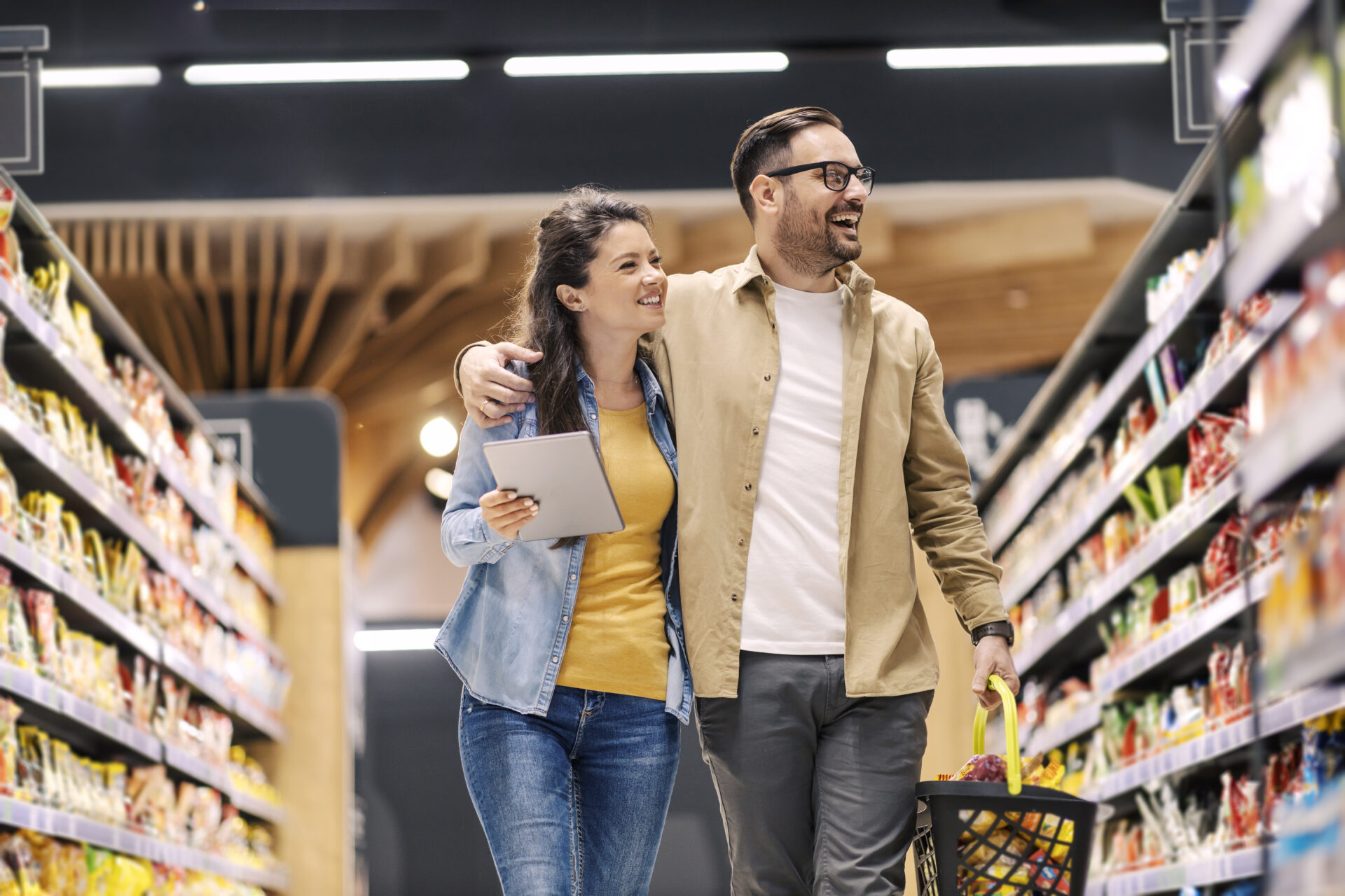 happy-couple-is-walking-trough-supermarket-carrying-list-with-groceries-tablet-scaled